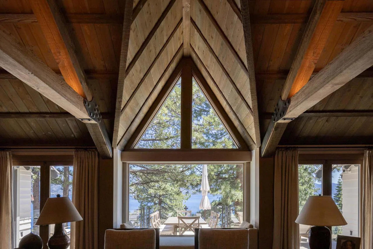 Lodge living room with vaulted wood ceiling and expansive triangular window framing outdoor views of trees and water.