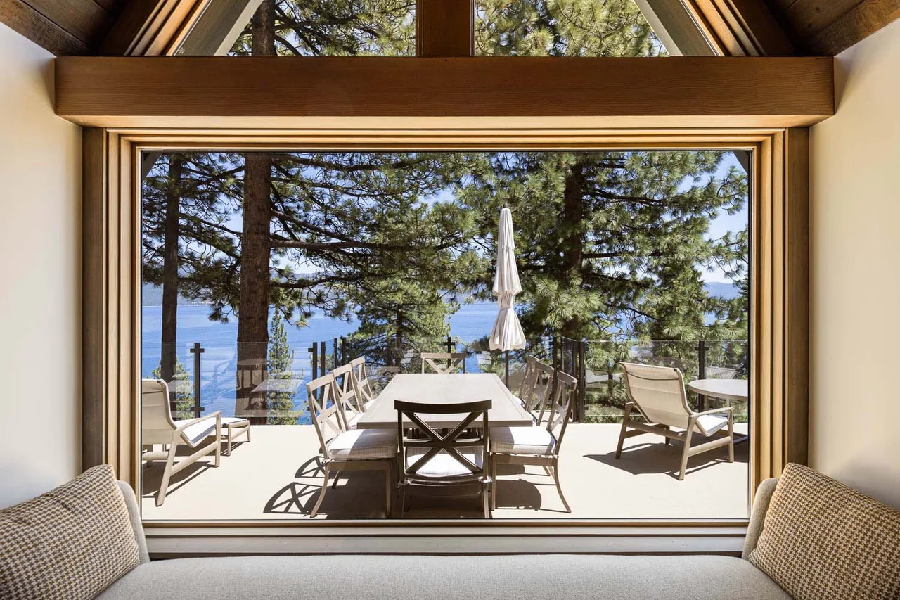Outdoor patio with dining set and umbrella, viewed through a large picture window framed in wood overlooking lake and trees.