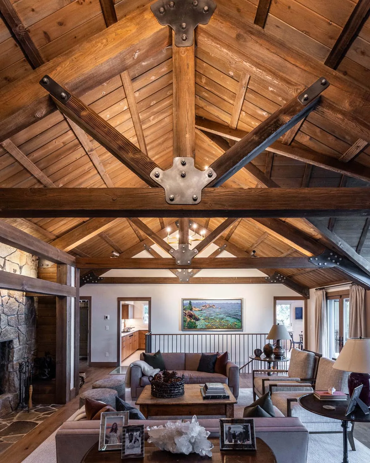 Living room features wood ceiling trusses, stone fireplace, and views toward a kitchen and outdoors.