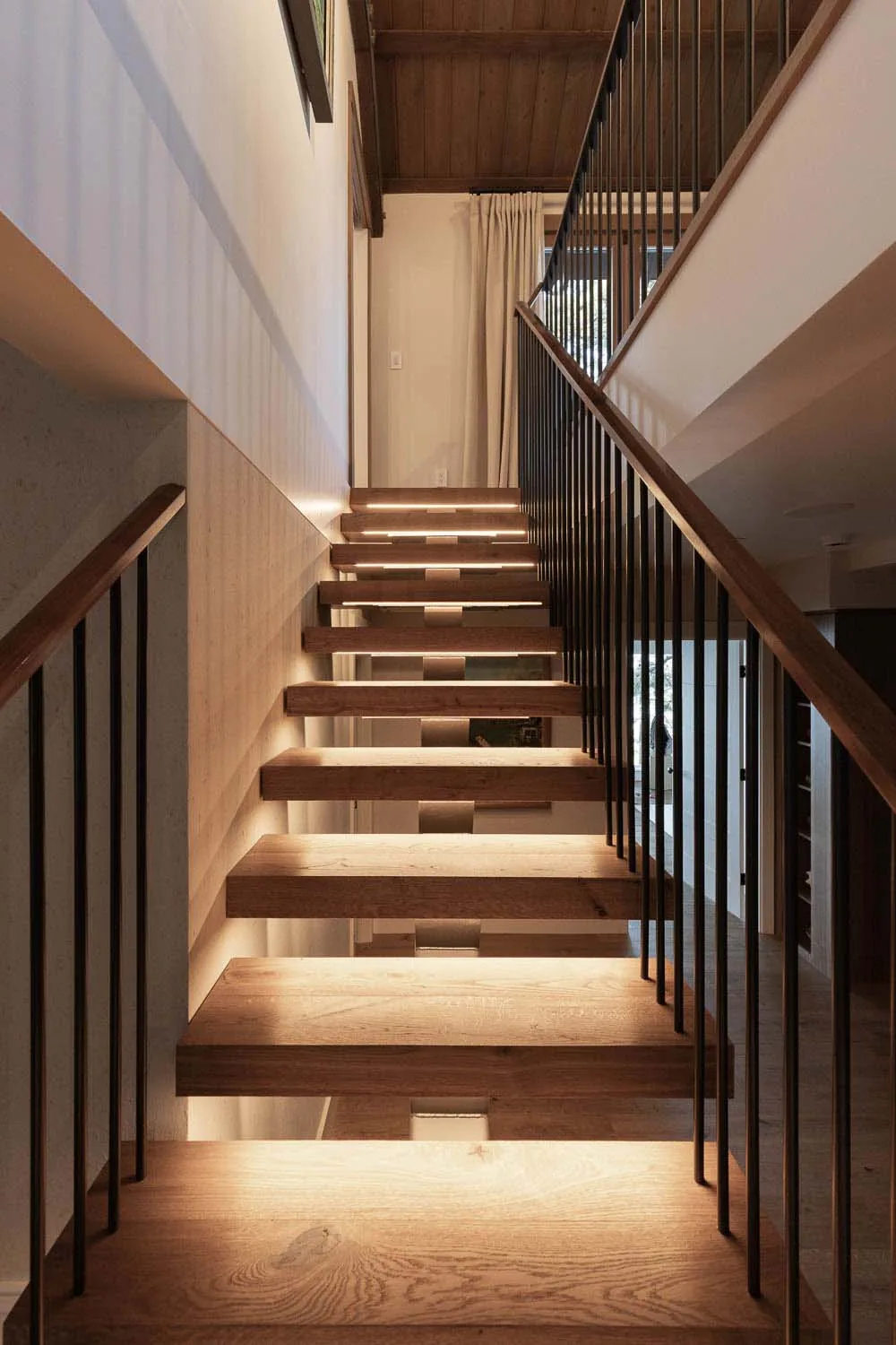 Floating wood stairs with integrated under-step lighting and black metal railings lead to an upper floor beneath a wood ceiling.