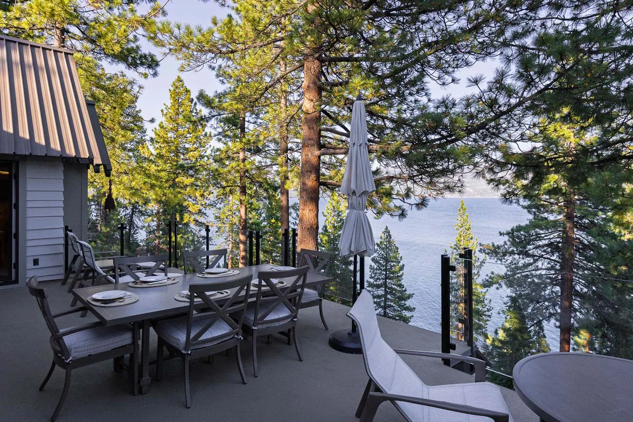 Outdoor dining area on patio with metal furniture and glass railing overlooking lake and trees.
