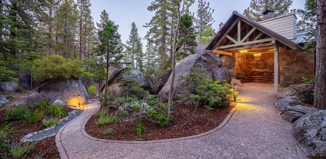 Pathway leads through a natural rocky landscape to a covered outdoor seating area with wood beams and stone walls.