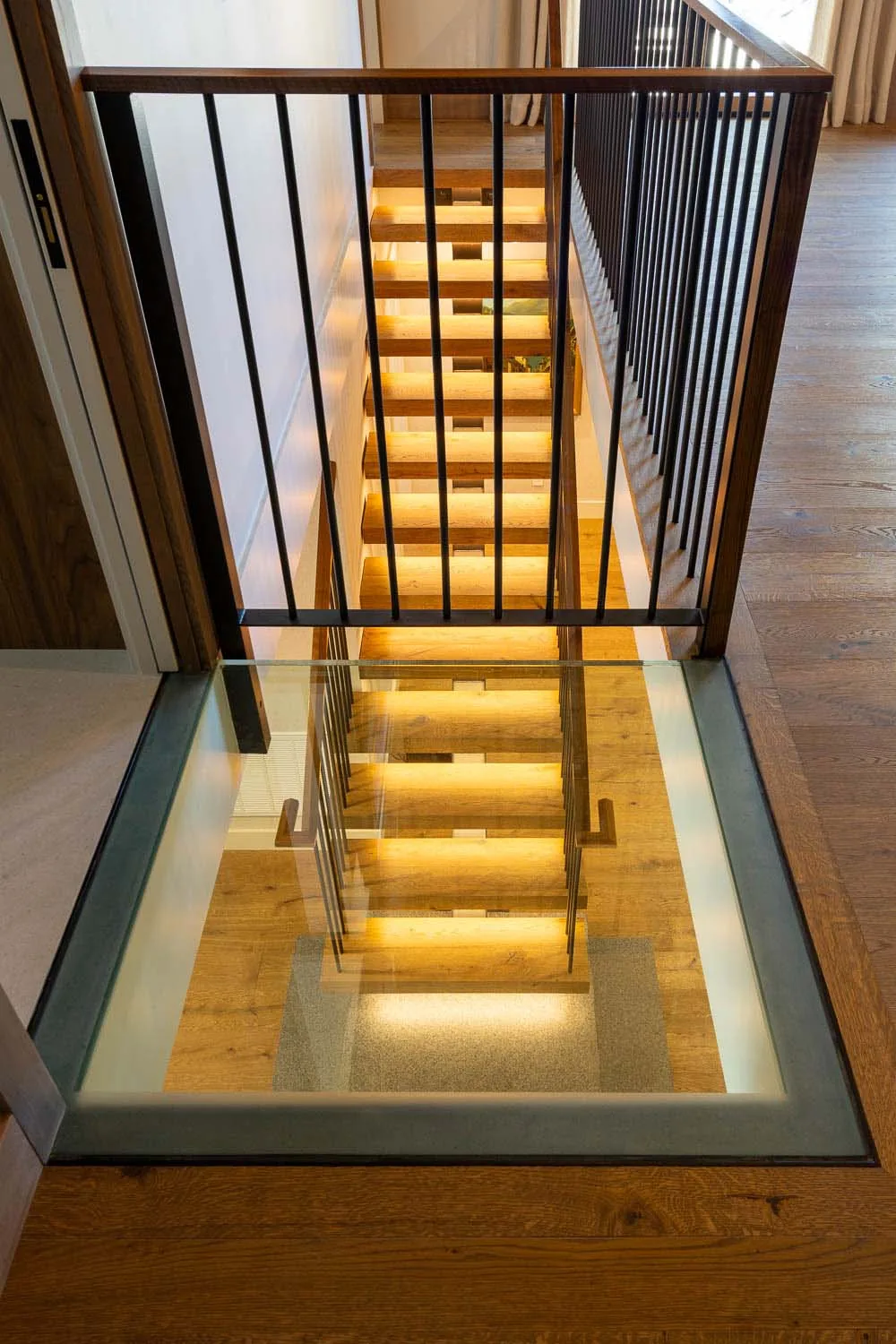 A staircase with wooden treads illuminated by under stair lighting is viewed through a glass floor panel with metal spindle railing above.
