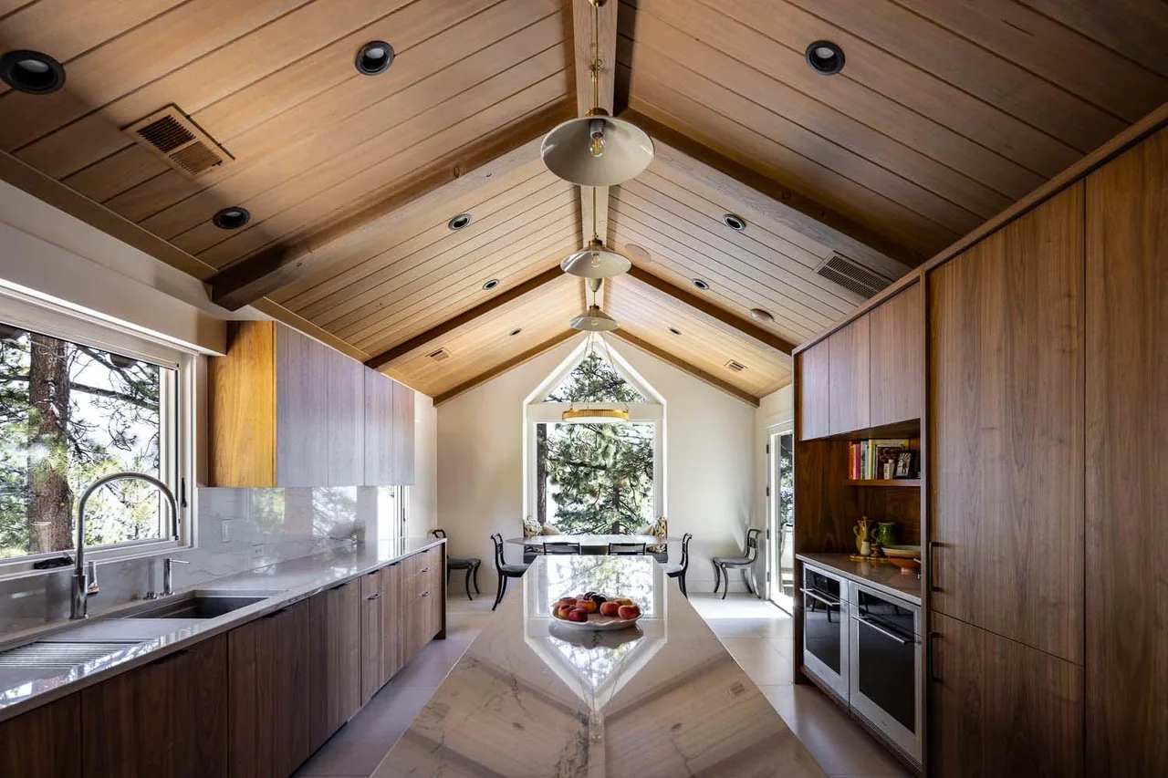 Kitchen with vaulted wood ceiling, marble island, and large window framing forest view in the Bold Tahoe Lodge Transformation.