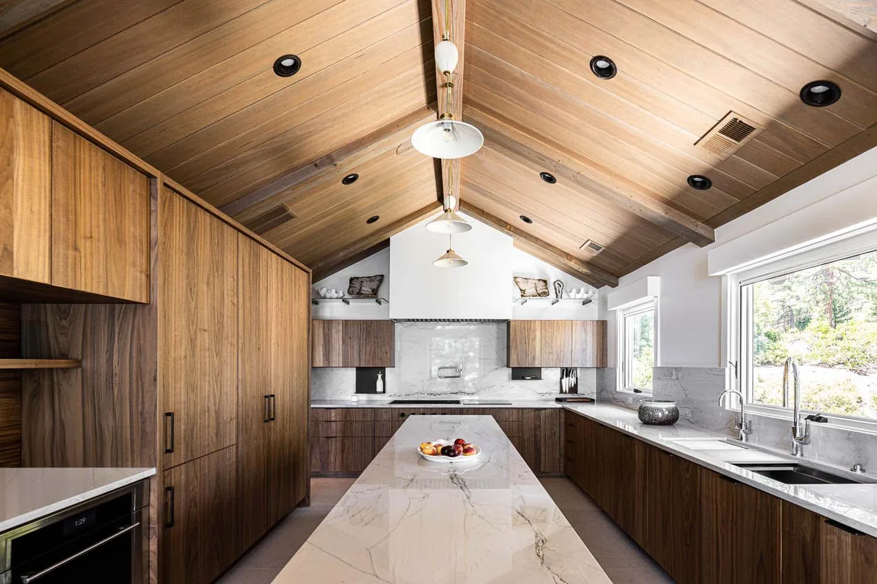 Kitchen features vaulted wooden ceiling, marble island, large windows, and dark wood cabinetry in the Bold Tahoe Lodge Transformation.