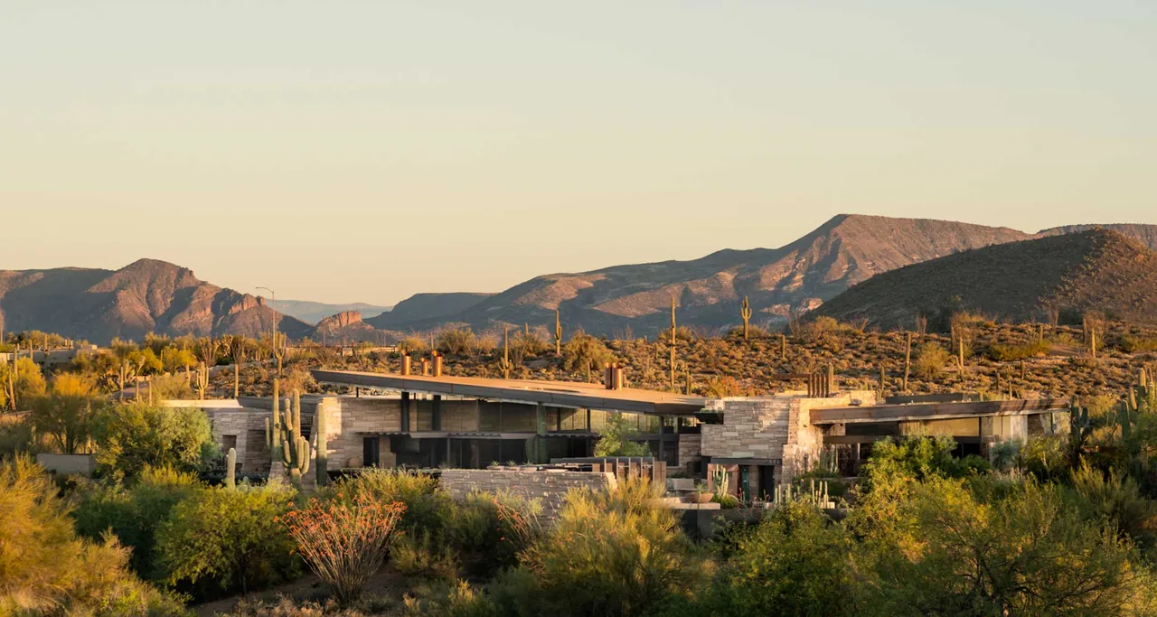 Saguaro Ridge features a modern desert home with stone cladding and a sloped roof, set against a mountain backdrop at sunset.