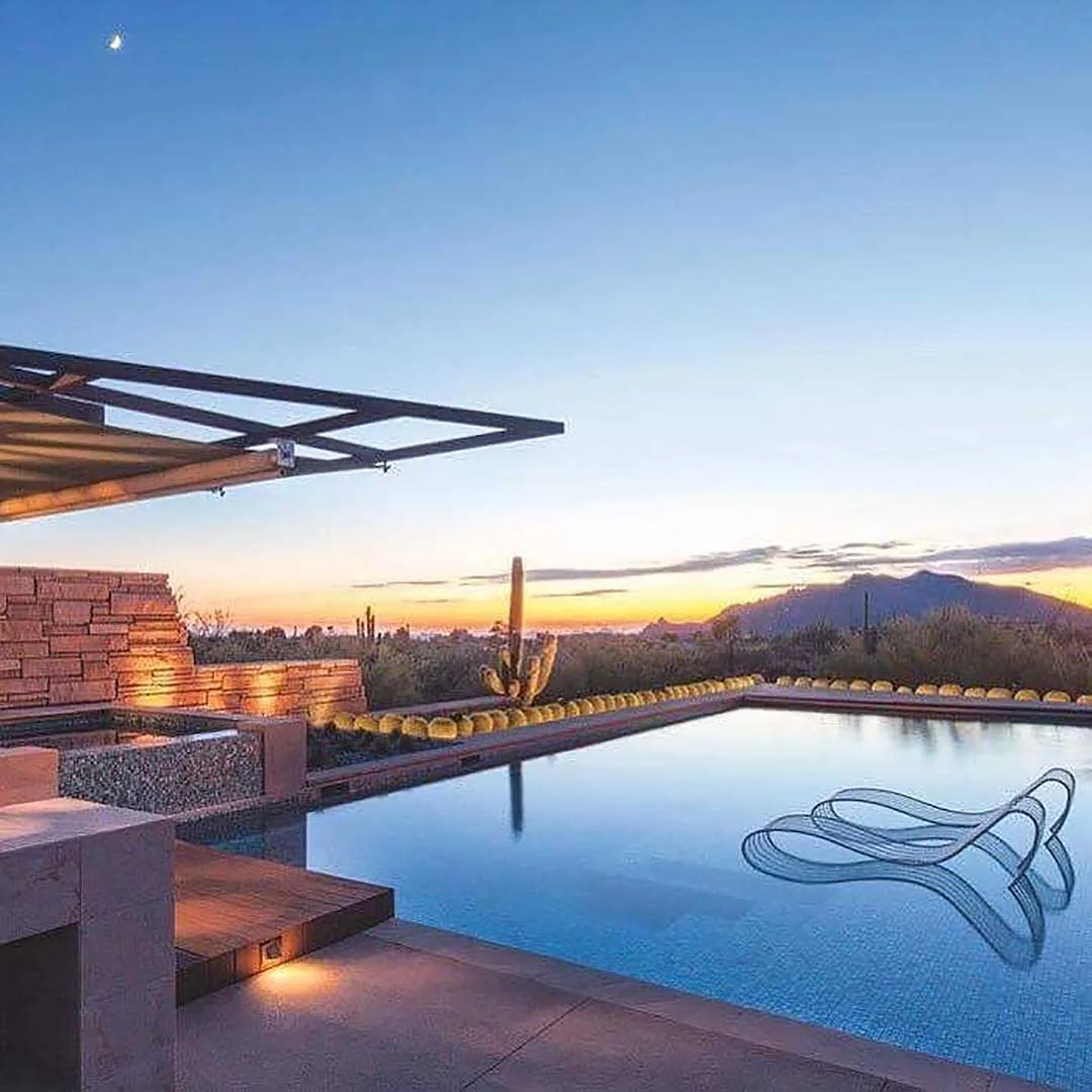 The Saguaro Ridge project features a pool with desert landscaping and a metal pergola in front of a mountain backdrop at sunset.