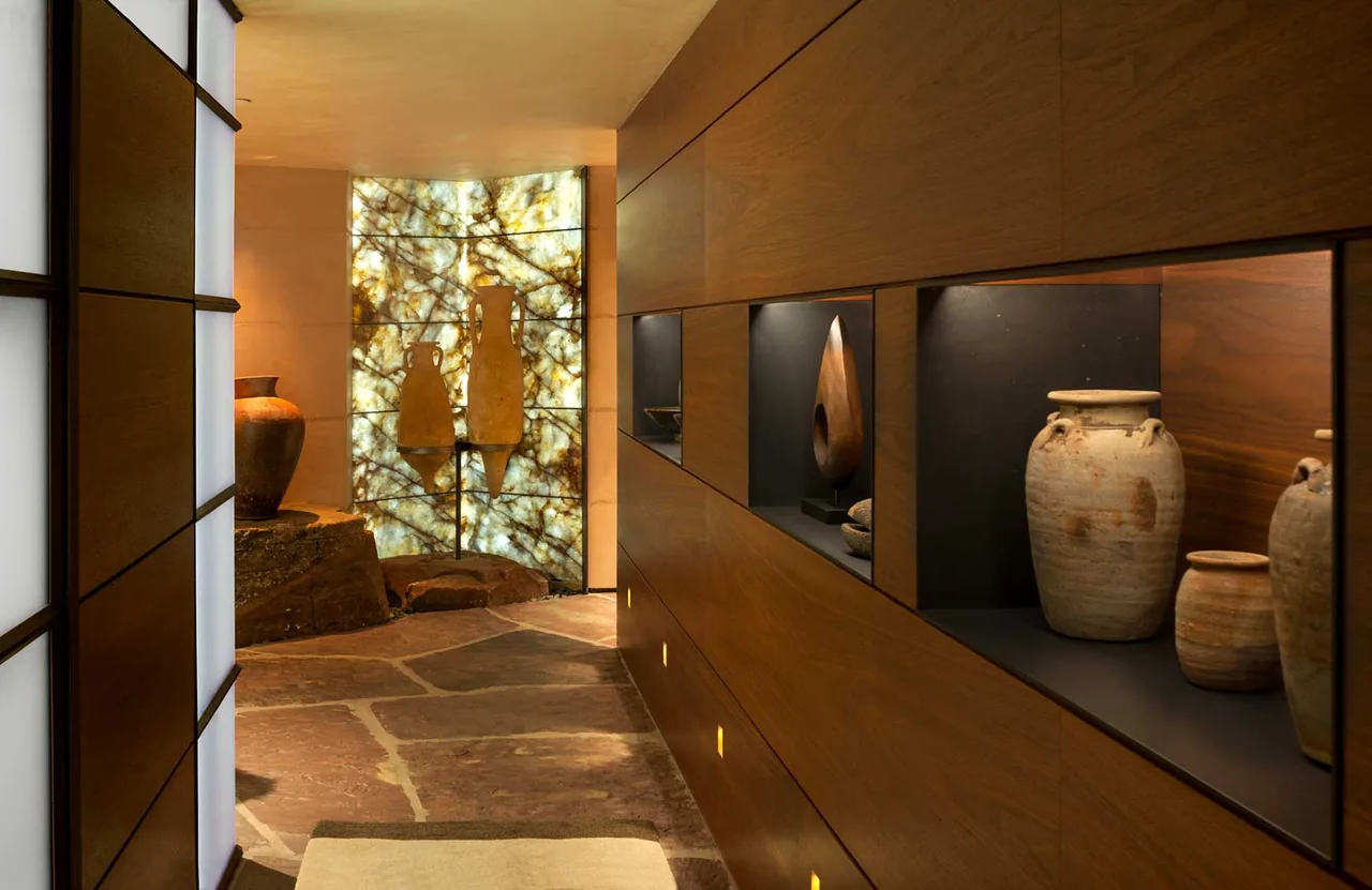 A hallway with wooden panel walls and built-in niches showcasing pottery, highlighted by a backlit decorative stone wall at the end.