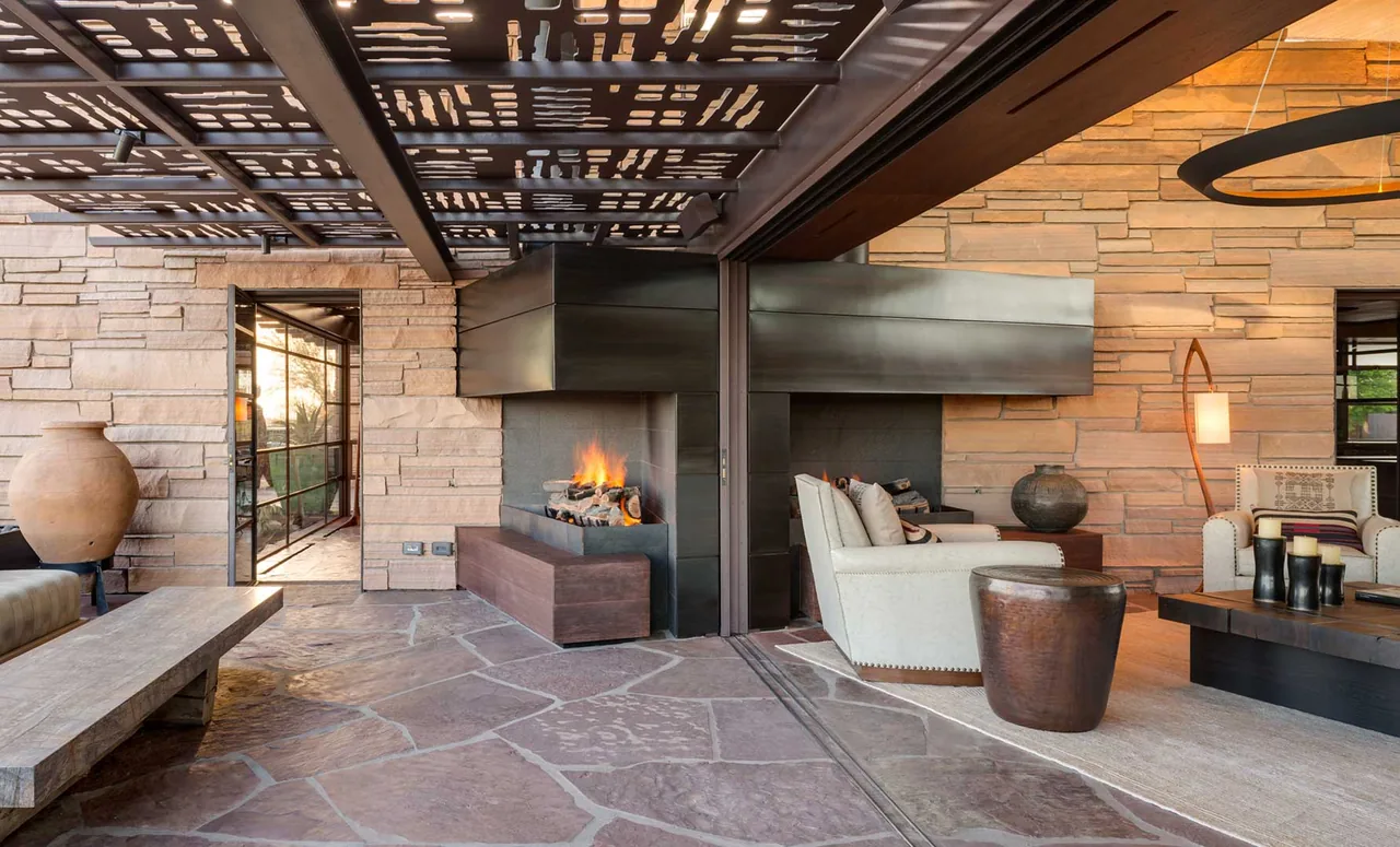 Covered patio features stone paving, a double-sided fireplace, and comfortable seating with desert-themed decor.