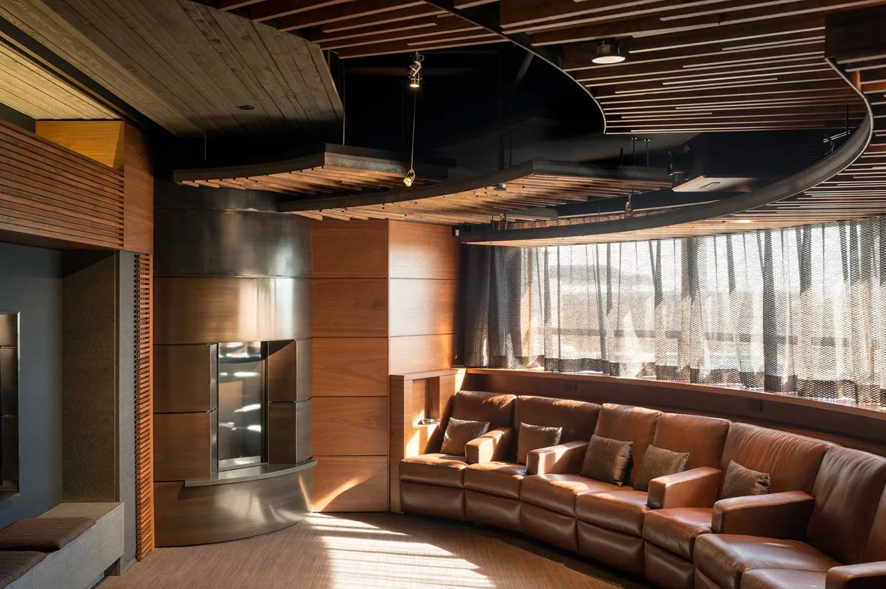 Living room with a curved leather sectional, wood panel walls, and a metal fireplace by Douglas Fredrikson Architects.