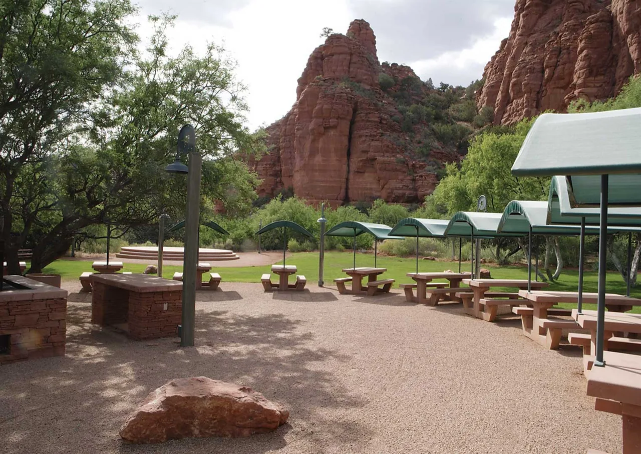 Covered picnic tables with curved metal roofs in an outdoor setting framed by red rock hillsides.