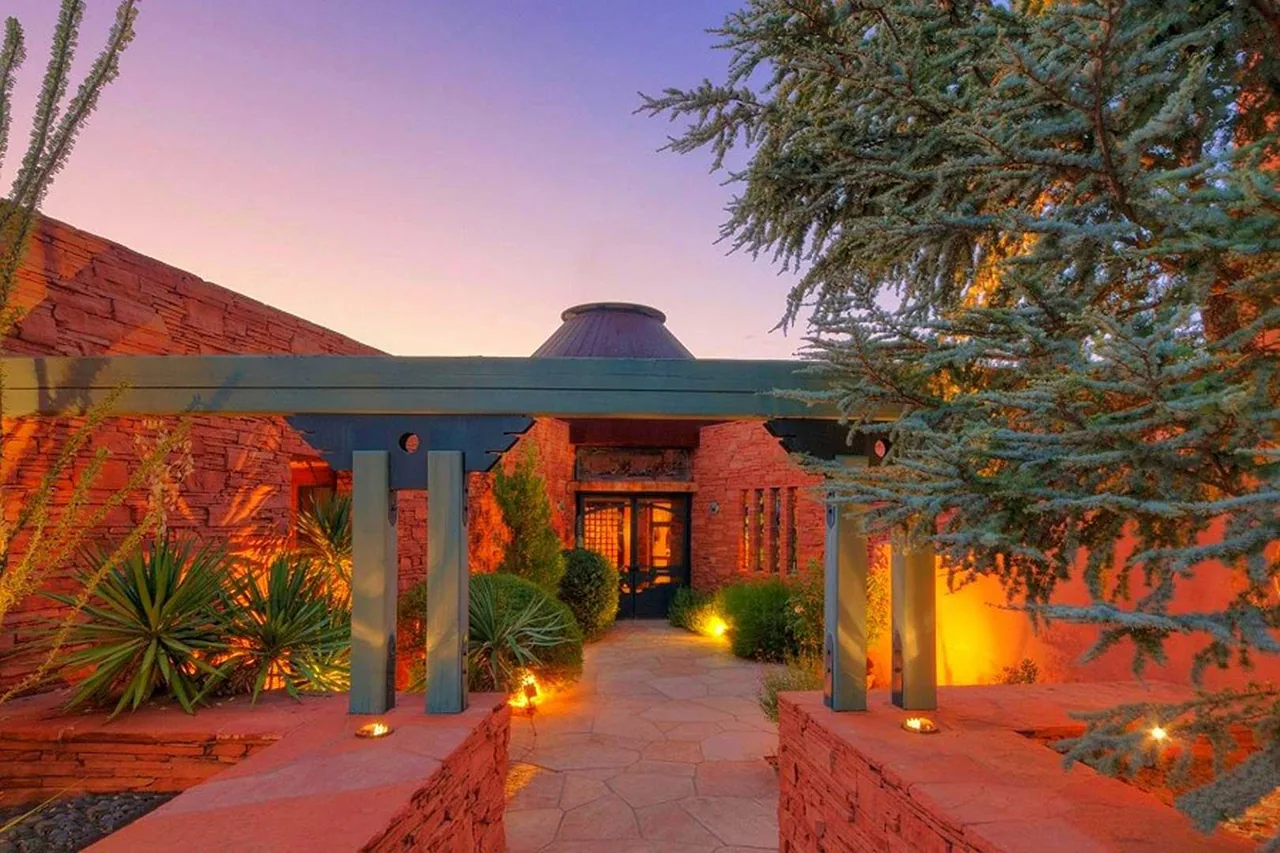 Red stone exterior walls and a wooden pergola frame the entrance pathway, lit by ground lights at dusk.