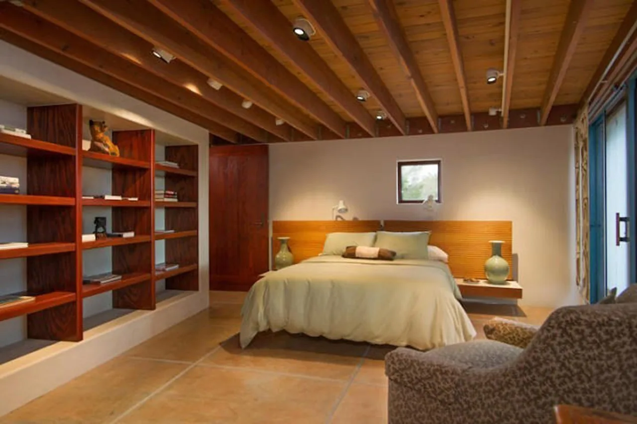 Bedroom featuring an exposed beam wood ceiling, built-in wooden shelves, and a bed with a horizontal slatted wooden headboard under a small square window.