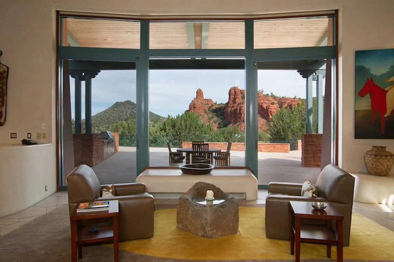 Living room interior with leather chairs, stone coffee table, and glass doors framing a desert landscape and patio seating.