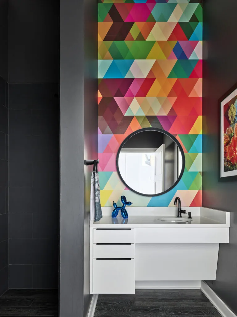 Bathroom vanity features a white floating cabinet under a round mirror with vibrant multicolored geometric wall art behind it.