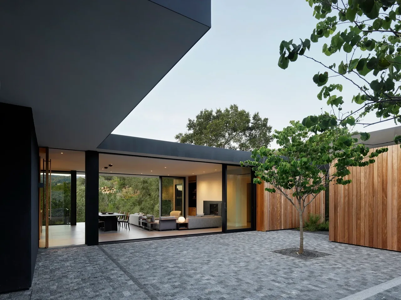 Modern home with an open courtyard featuring a single tree, wood siding, and floor-to-ceiling glass walls revealing living and dining areas.