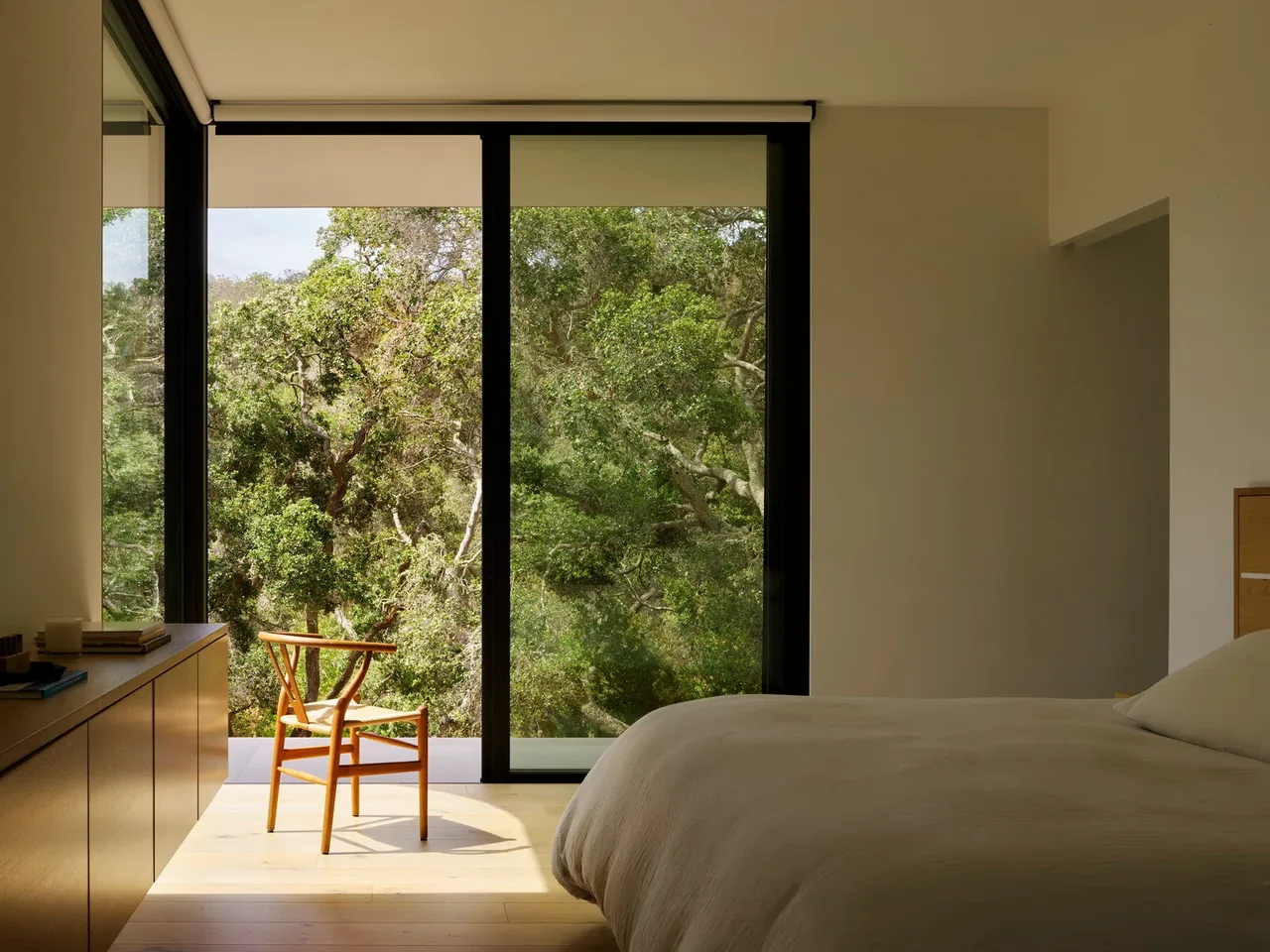 Minimalist bedroom features a large black-framed sliding glass door opening to a wooded view outside.