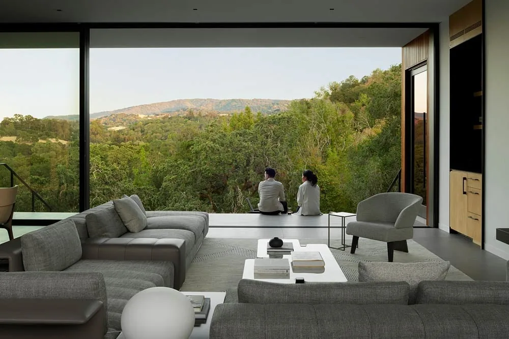Modern living room features large floor-to-ceiling windows with an expansive view of a forested hillside.