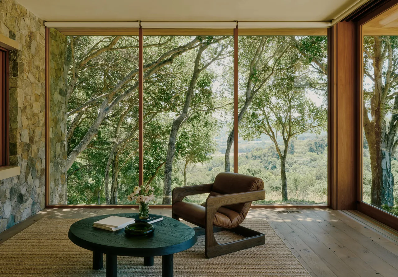 A seating area with a leather armchair and round table framed by floor-to-ceiling glazing and a stone wall.