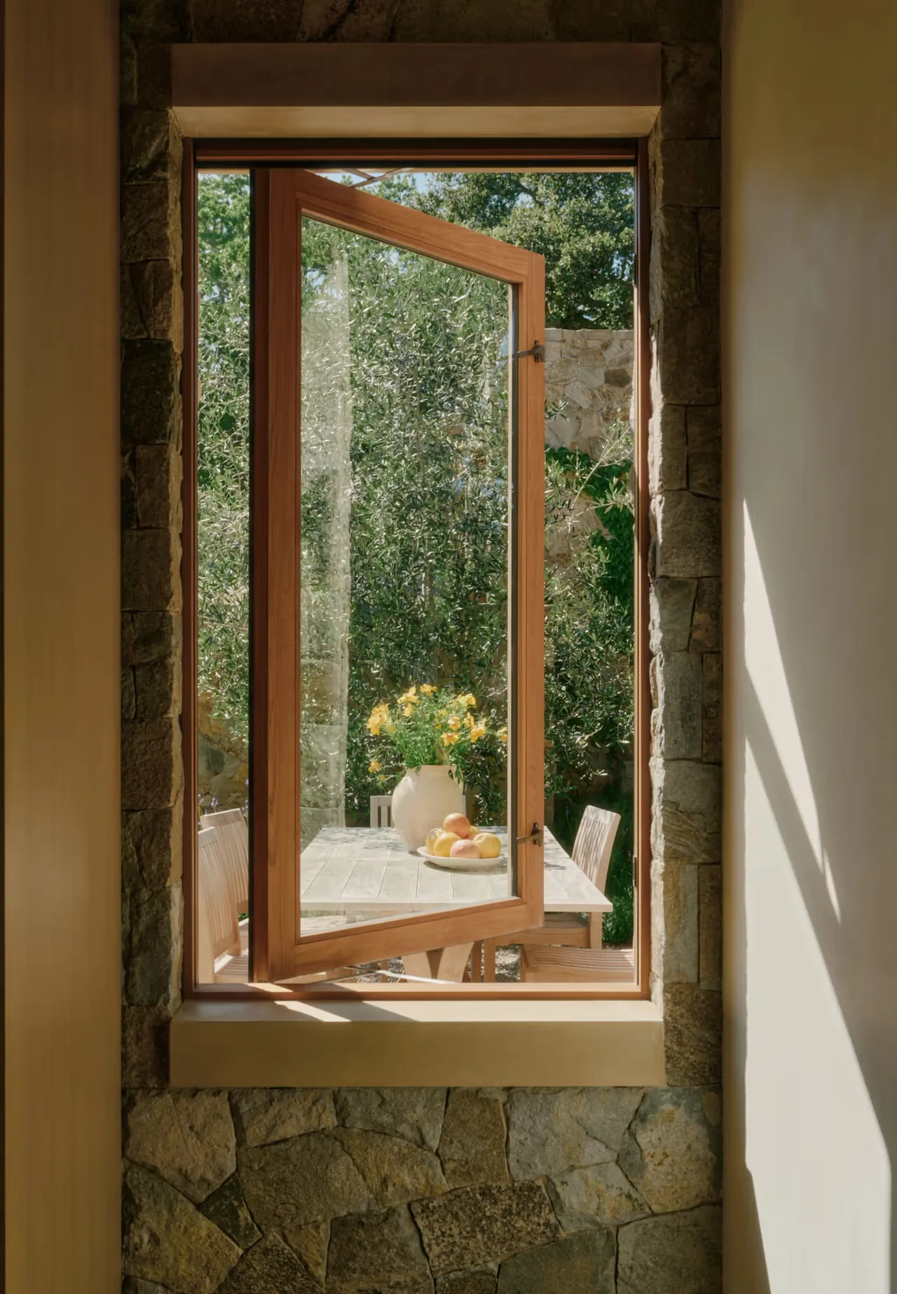 Open wood-framed casement window reveals a shaded outdoor dining area with natural stone walls and garden greenery.