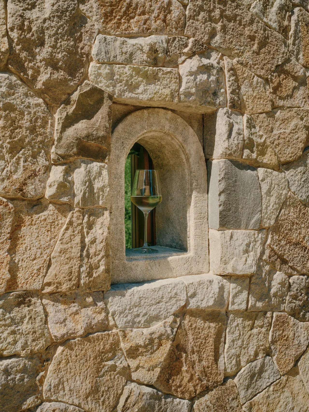 Detail of a natural stone wall featuring a carved arched niche with a glass of white wine inside.