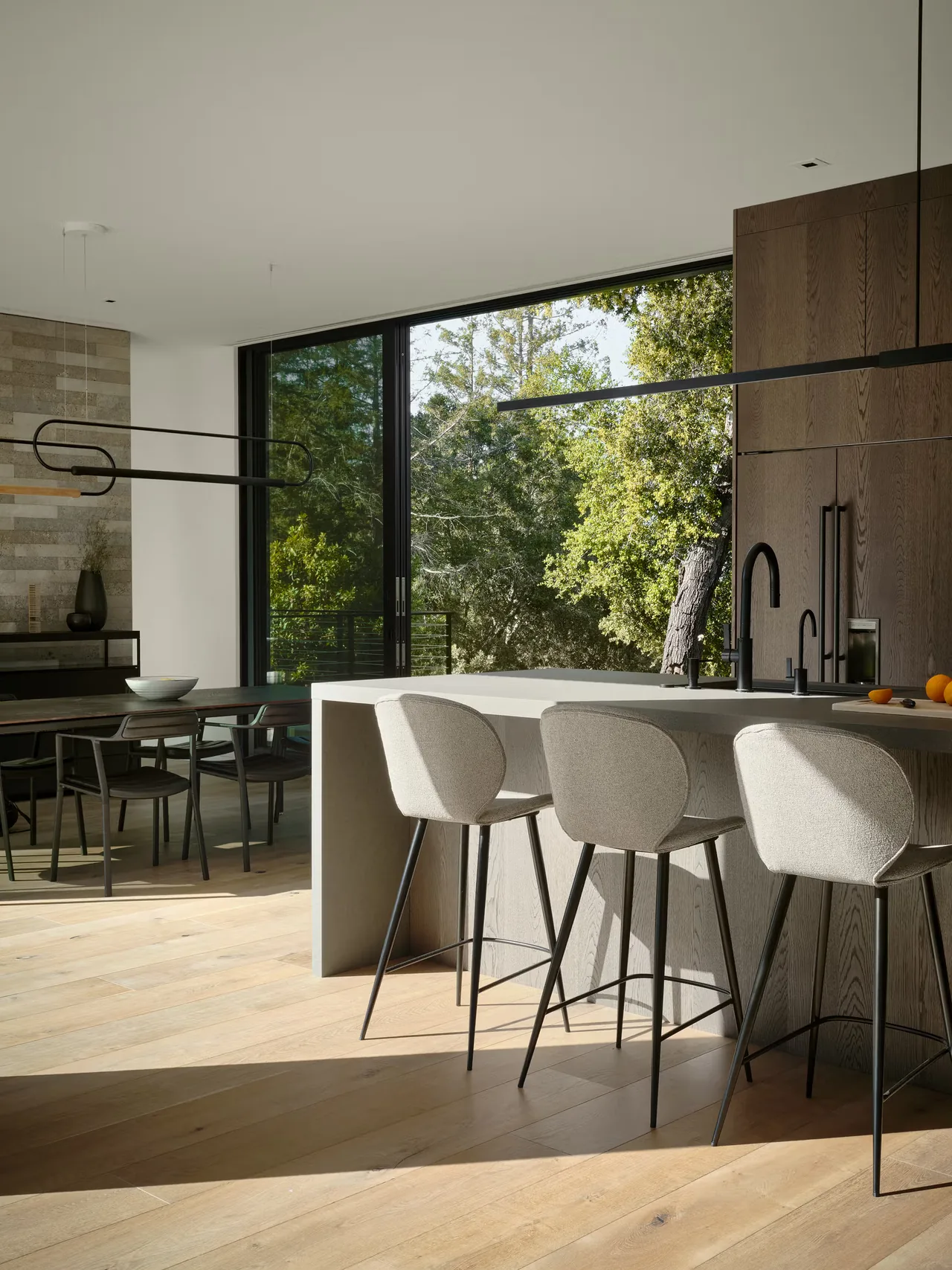The Guzhai kitchen features a light wood floor, a minimalist island with gray bar stools, and expansive sliding doors to a green outdoor view.