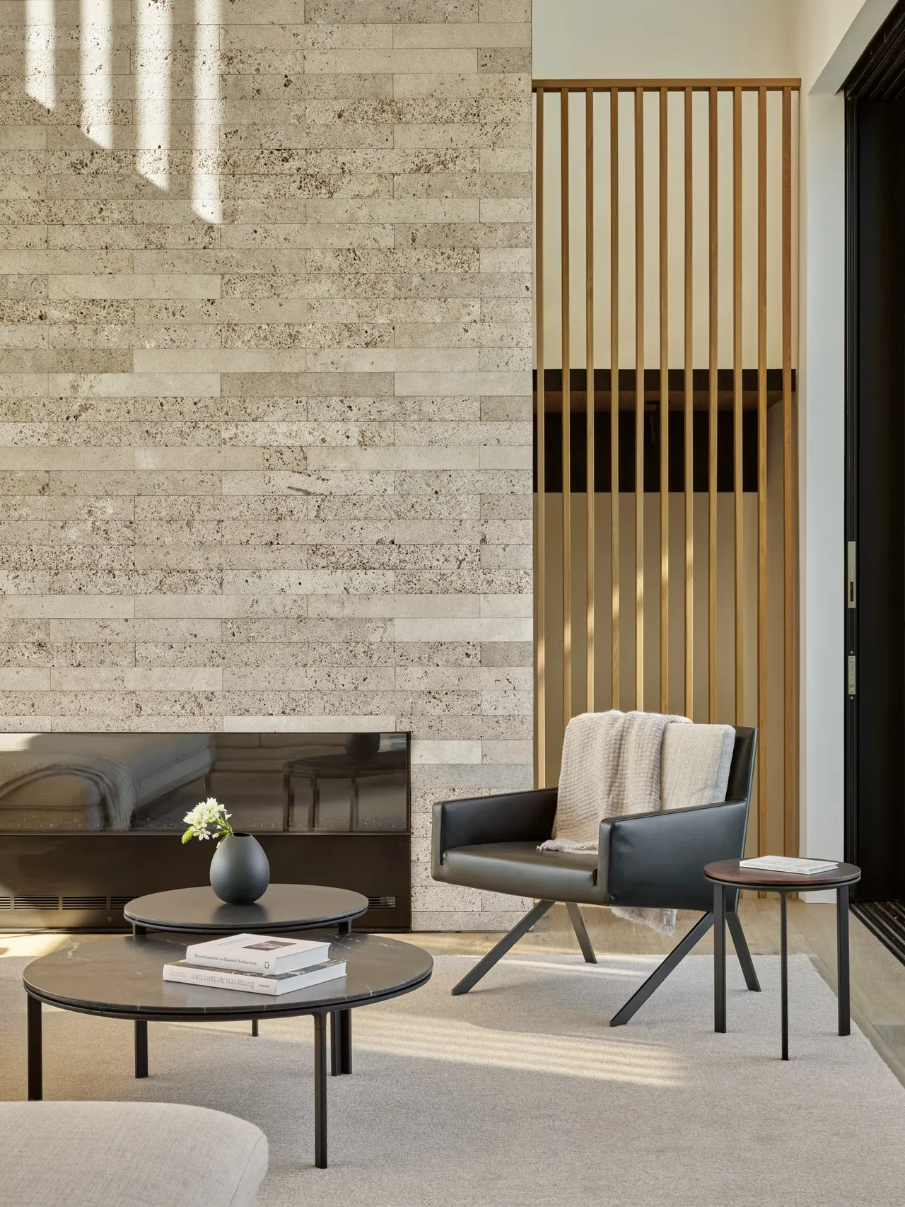 Living room corner features a textured stone wall, black leather chair, and nested round coffee tables with books and a vase.