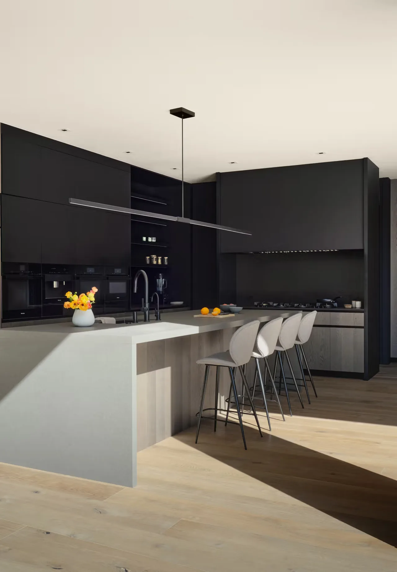 A spacious, modern kitchen with a light gray island, black cabinets, and bar seating for four under a sleek linear pendant light.