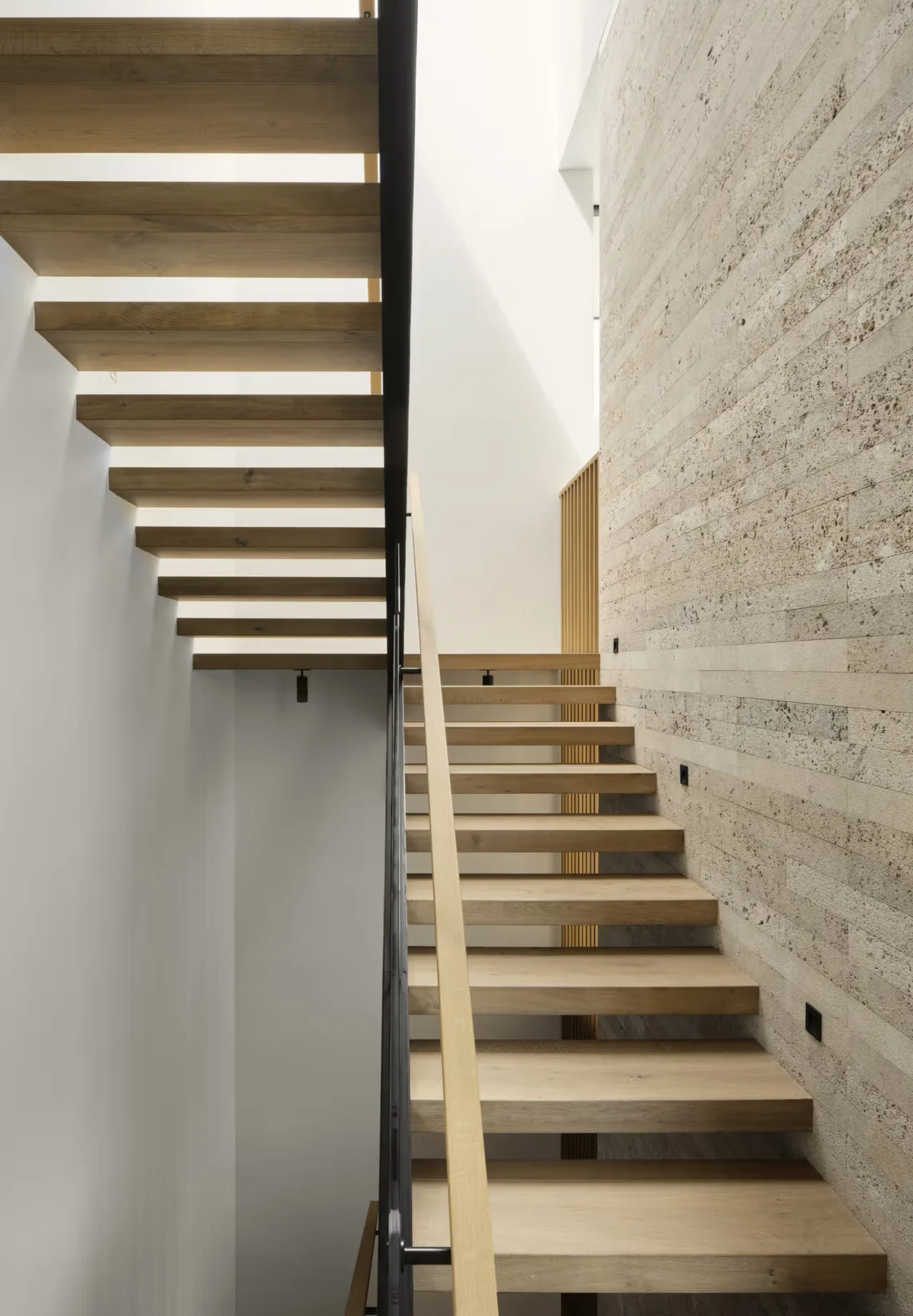 Floating wood staircase with open risers and a textured stone wall in Guzhai project by Feldman Architecture.