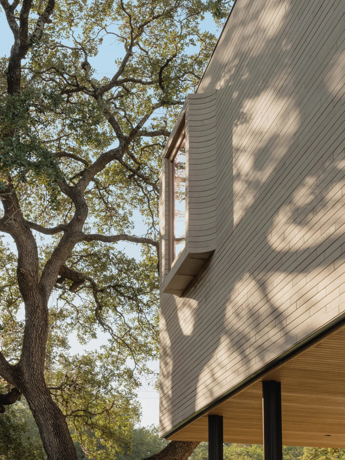 Greenbelt Residence features light brick cladding with a large vertical window and subtle shadow patterns from nearby trees.