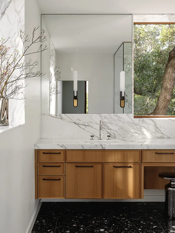 Bathroom vanity with marble countertop, wood cabinets, and a large mirror, featuring a view of a tree through a window.