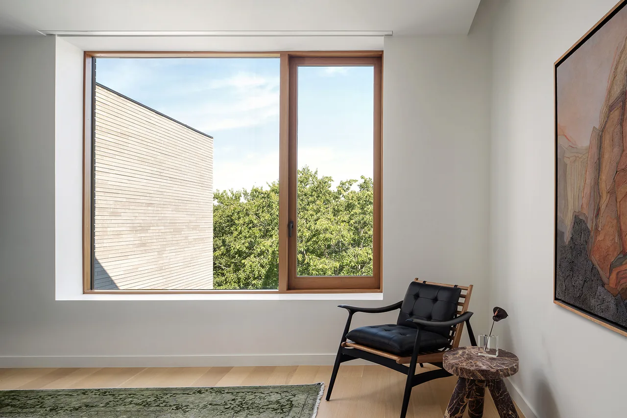 Interior corner with a tall vertical window framing green trees, featuring a wood chair with black cushions and a marble side table.