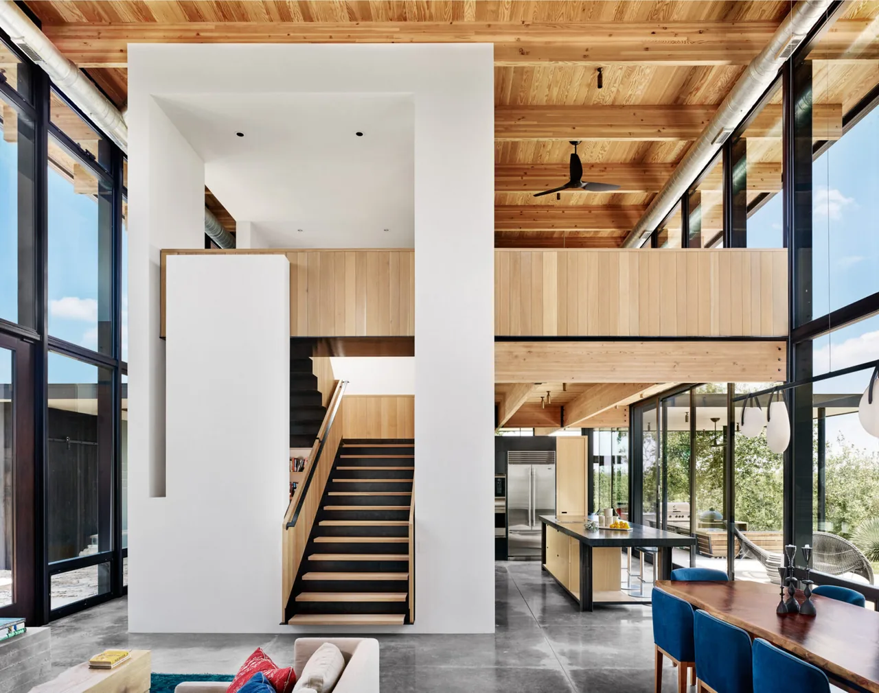 Modern interior with black window frames, exposed wooden ceiling beams, and an open stairway leading to a mezzanine level in Llano Retreat.