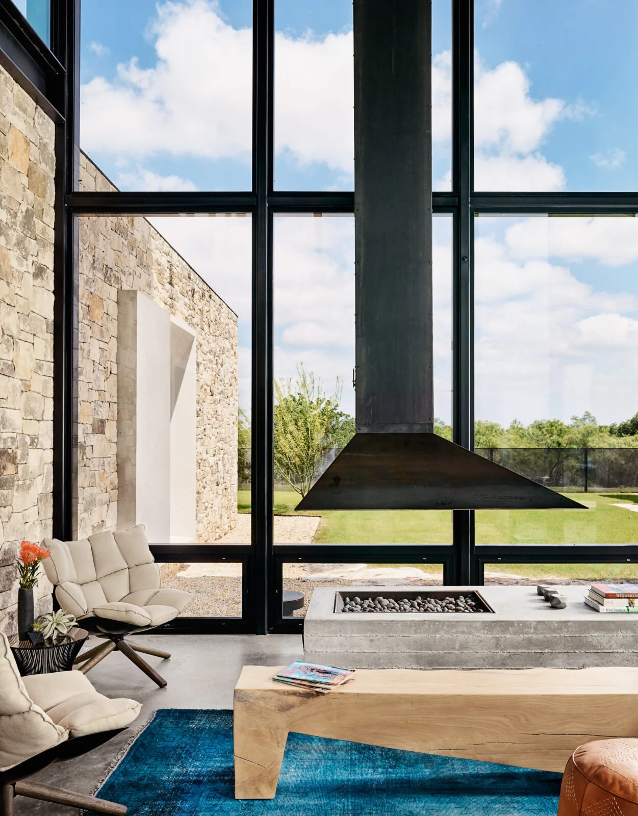 The Llano Retreat living area features floor-to-ceiling black framed windows with a suspended metal fireplace hood and a textured stone exterior wall.