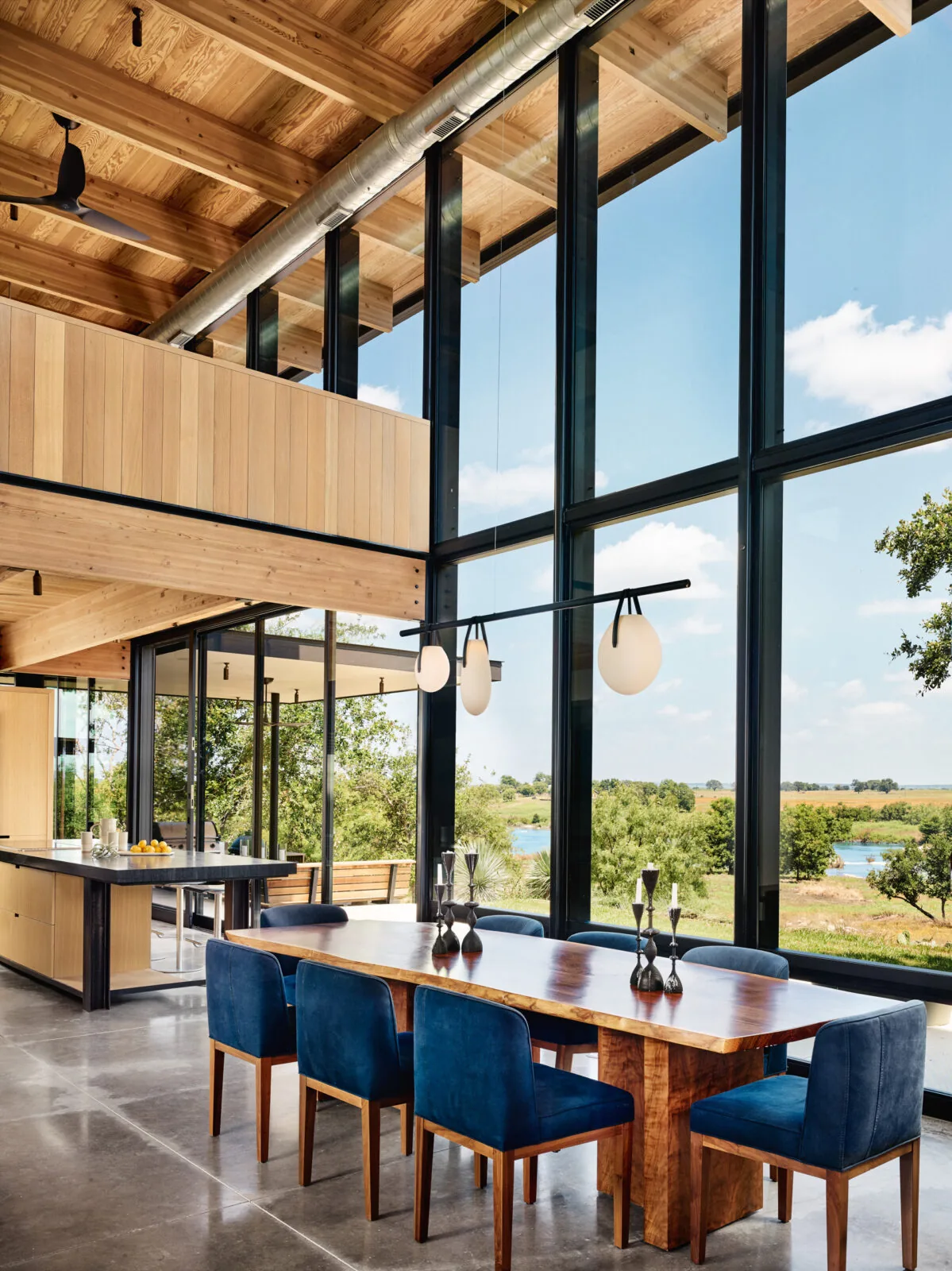 Dining space with floor-to-ceiling glazing and blue velvet chairs at a wooden table, showcasing wood ceiling beams and polished concrete flooring.