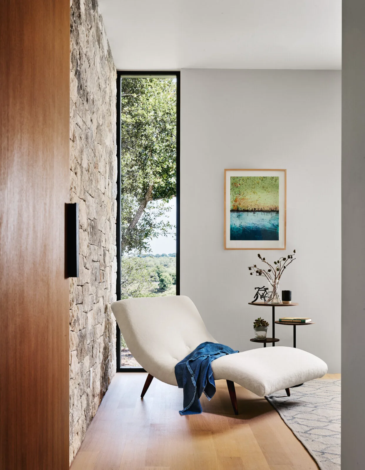 Curved white chaise lounge by a floor-to-ceiling window with views of greenery and a stacked stone wall.
