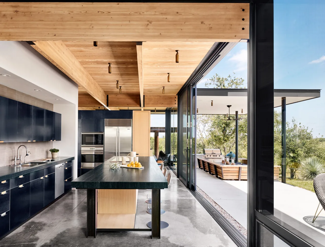 Modern kitchen features dark cabinets, concrete flooring, and large island opening to a covered outdoor living patio.