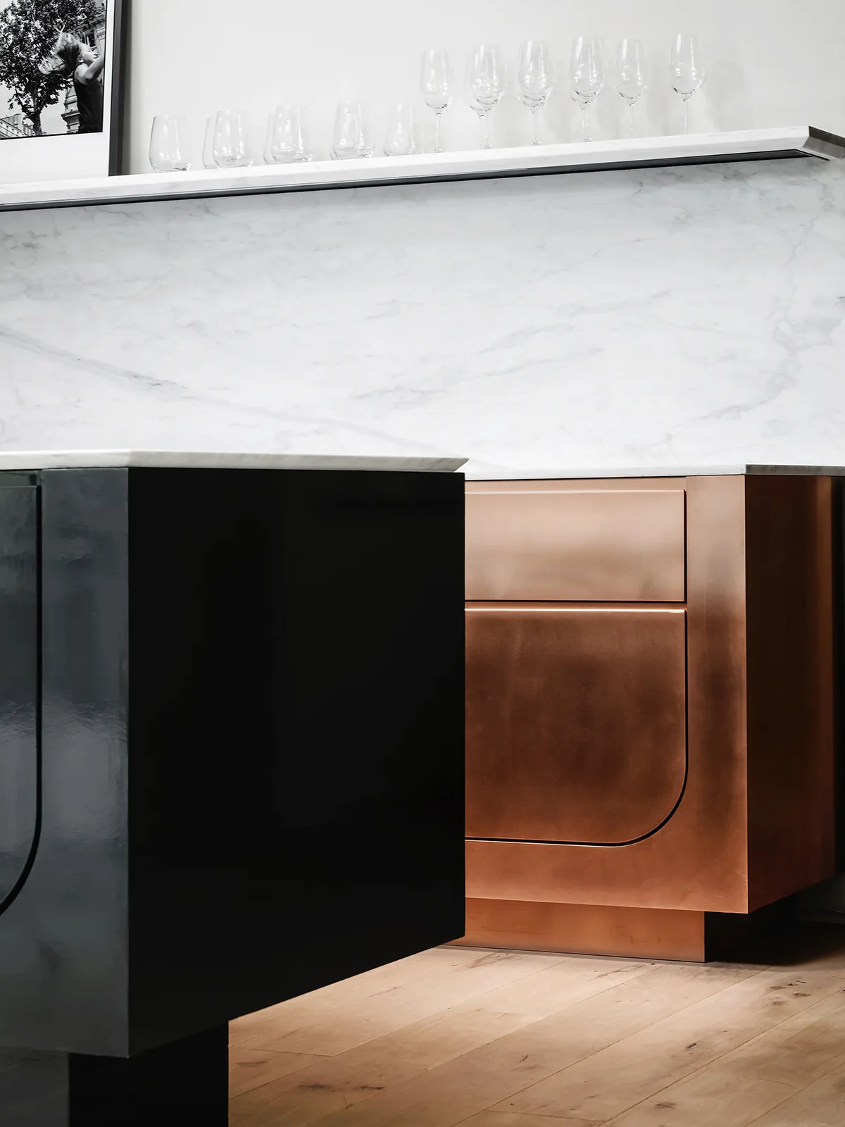 Detail of kitchen cabinetry featuring black and copper finishes with marble backsplash and glassware on a floating shelf.