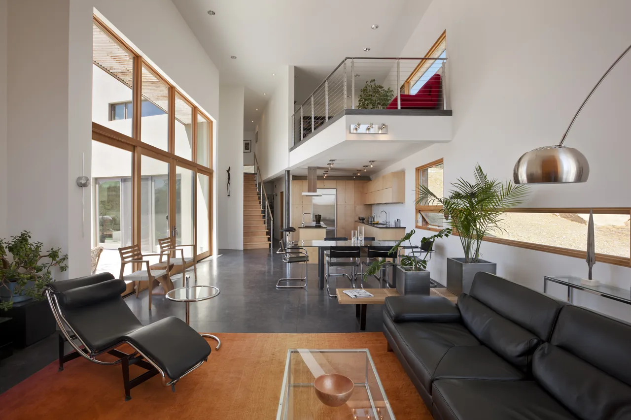 Living area with open kitchen and dining, featuring floor-to-ceiling windows and a mezzanine overlooking the space.