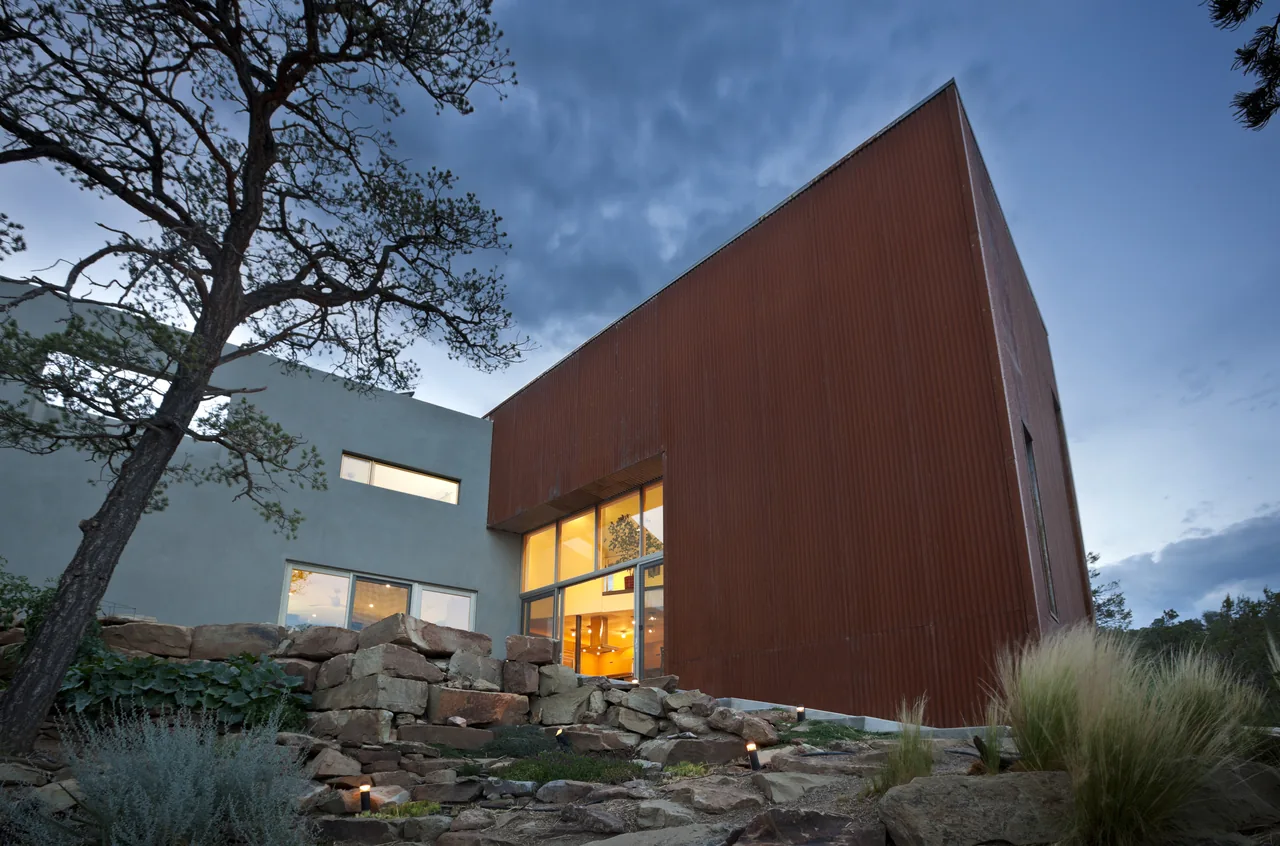Angular modern house with rusty metal siding and large windows seen from a rocky landscaped yard at dusk.