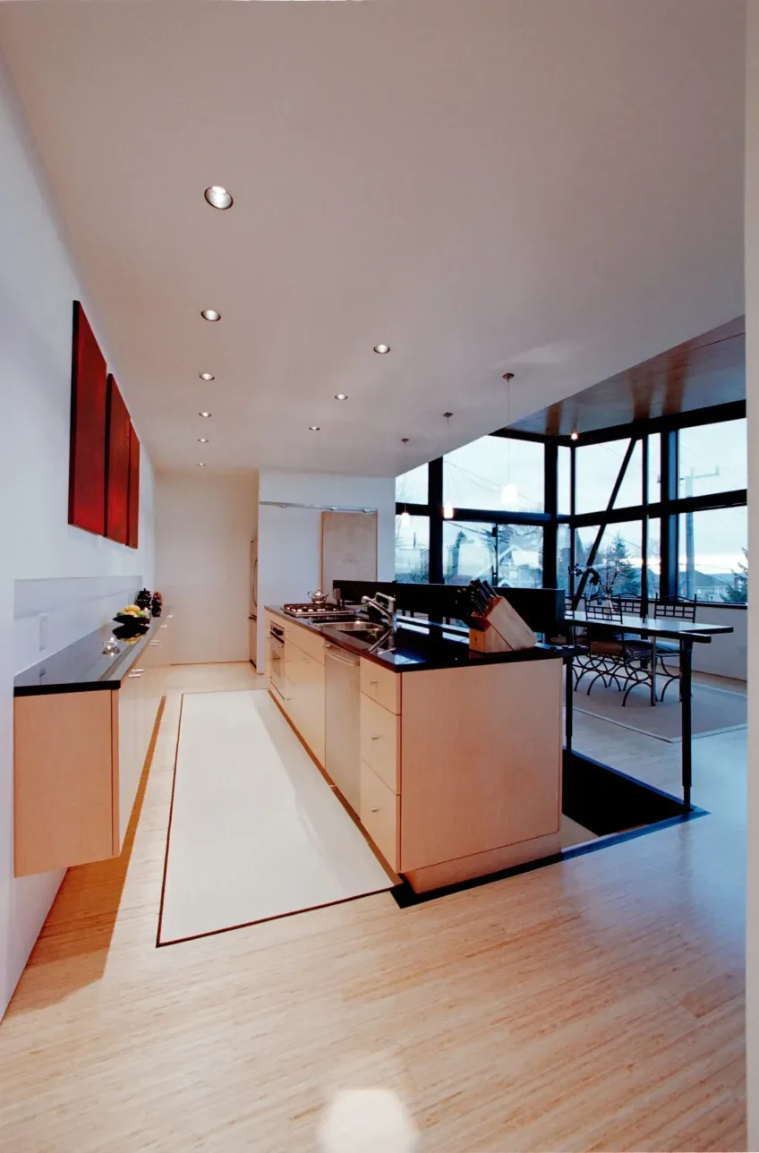 Modern kitchen featuring light wood cabinets, black countertops, recessed and pendant lighting, with large windows adjacent to the dining space.