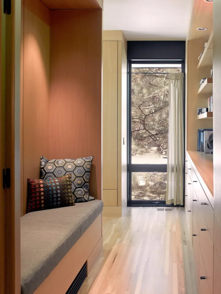 A narrow space features a built-in cushioned bench with pillows, continuous wood cabinetry, and a floor-to-ceiling window with a light curtain.