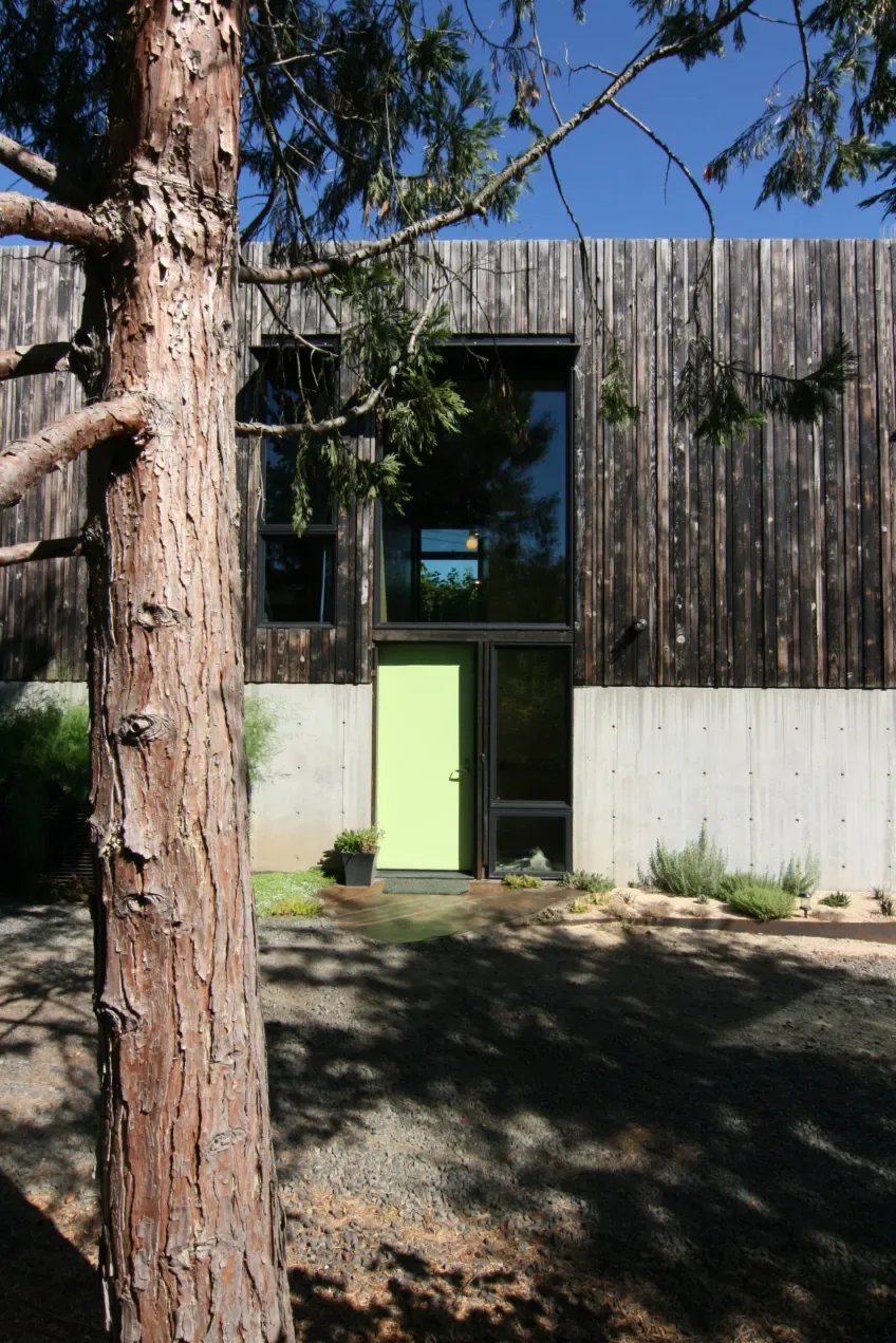 Modern exterior of House for a Cabinetmaker with weathered wood siding, concrete base, and black window frames.