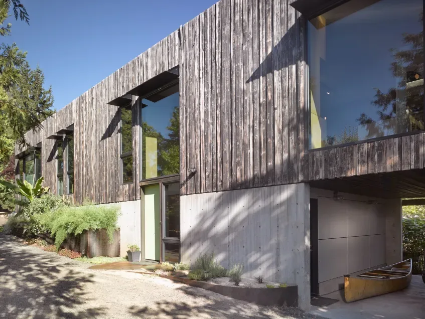 House for a Cabinetmaker features vertical weathered wood siding above a concrete base with large windows and a garage below.