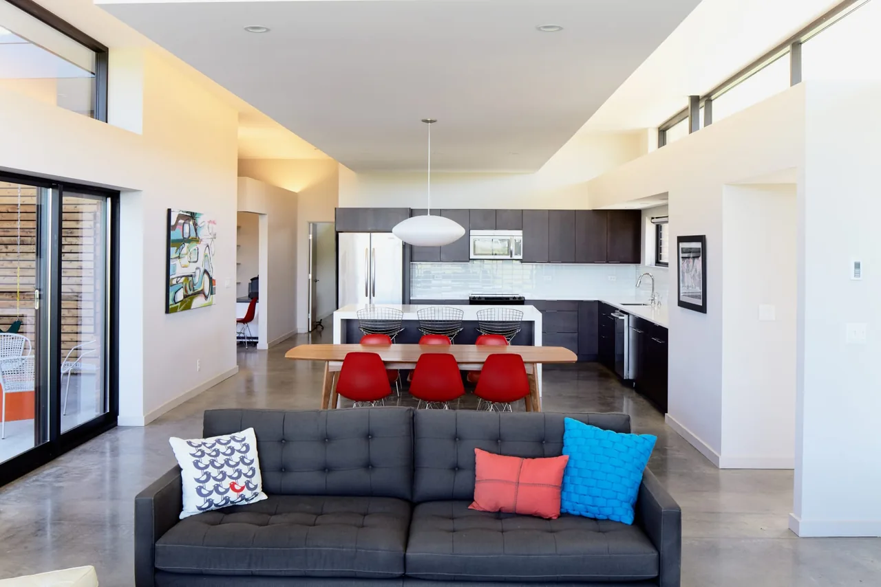 Contemporary open living space featuring a kitchen with dark cabinetry, a wooden dining table, and colorful cushions on a gray sofa.