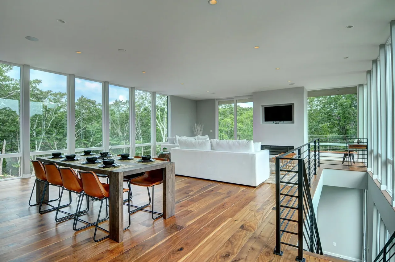 Open living and dining area with large windows, white sofa, wood floors, and a metal staircase railing.