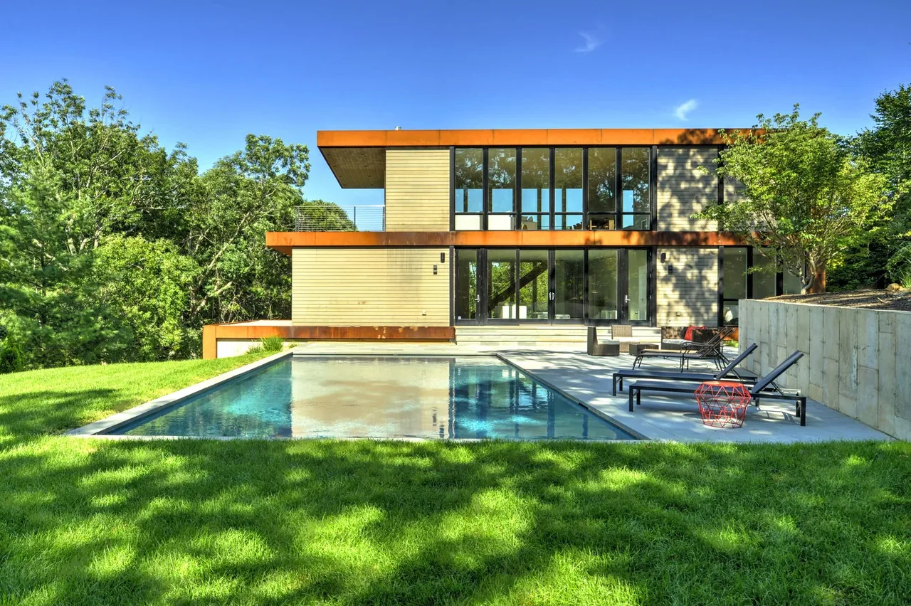Exterior rear view of a modern home featuring large glass windows and rust-colored steel detailing above a pool patio area.