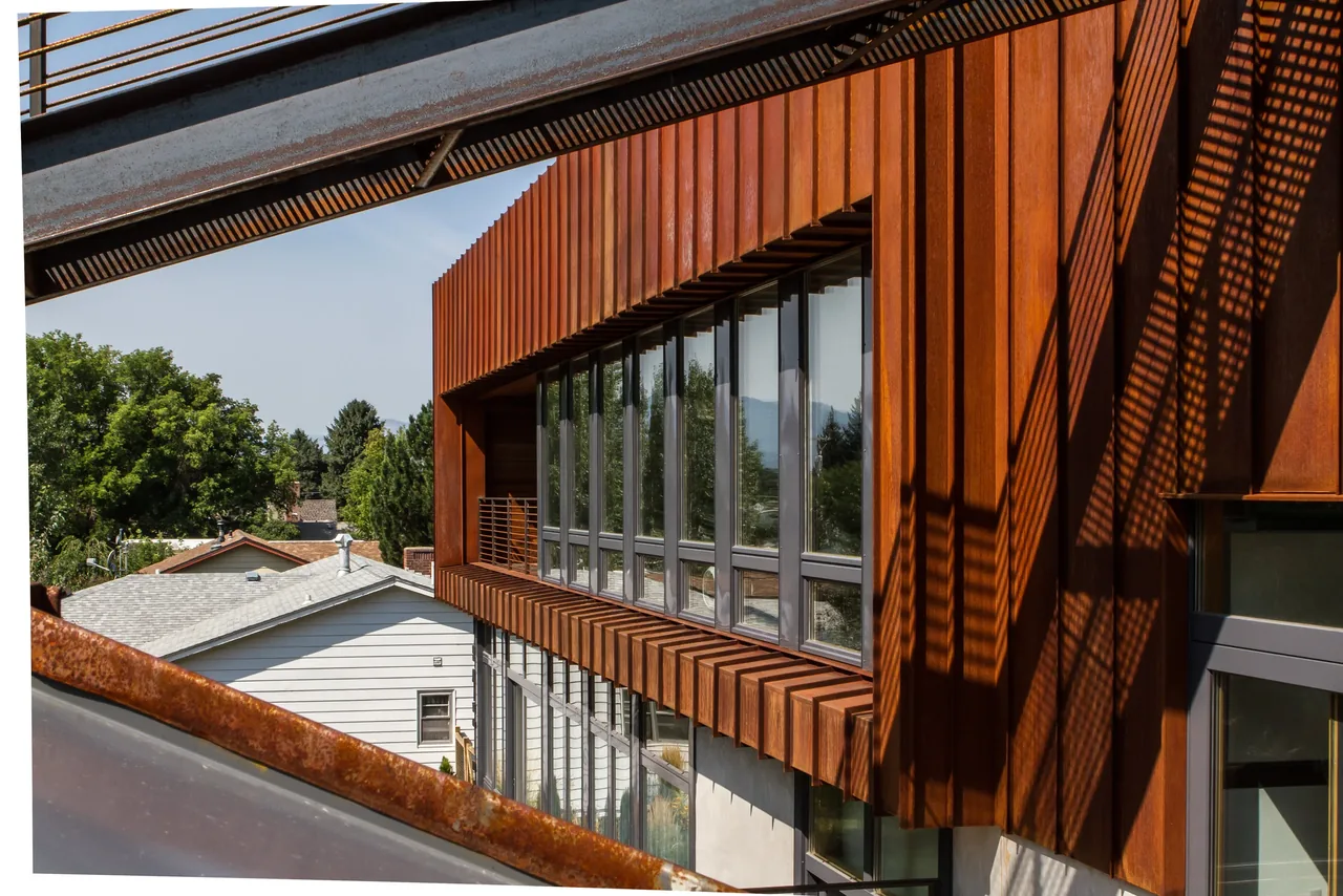 The Wren Residence features rust-colored metal siding and large windows on its upper floor with a suburban neighborhood backdrop.