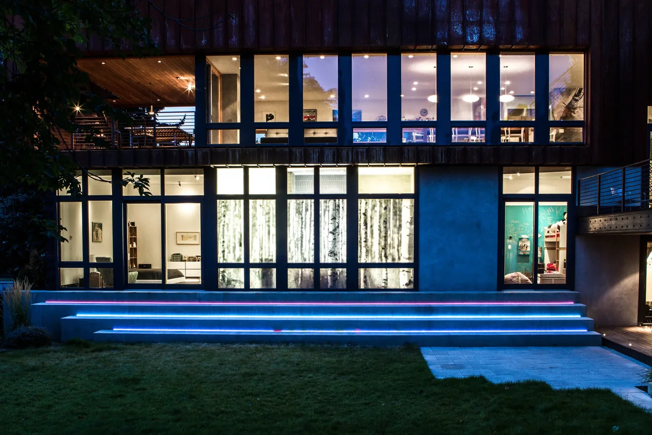 Wren Residence features large grid windows over illuminated concrete steps leading to a landscaped lawn in suburban Seattle.