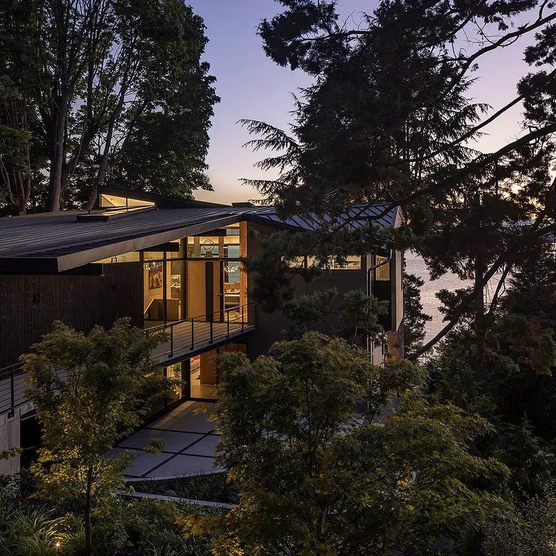 Modern home with sloped metal roof, floor-to-ceiling glazing, and wood railing at dusk overlooking water.